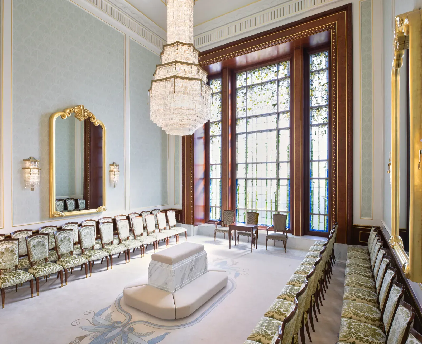 Sealing room with a central marble altar and cushioned bench, rows of upholstered chairs, gold-framed mirrors and tall leaf-patterned windows