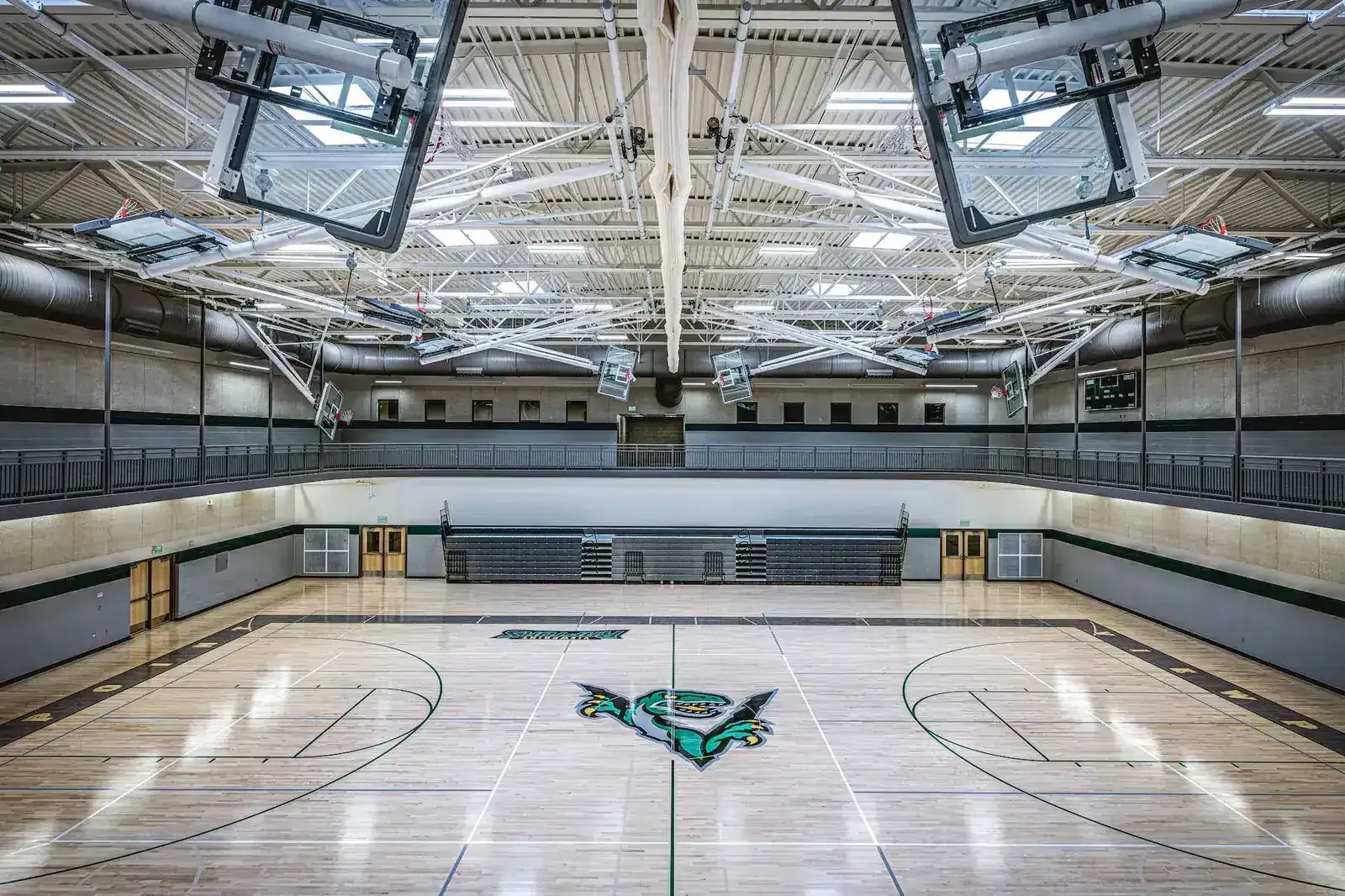 Gymnasium with a wood floor, a green mascot logo at center court, and an upper-level balcony.