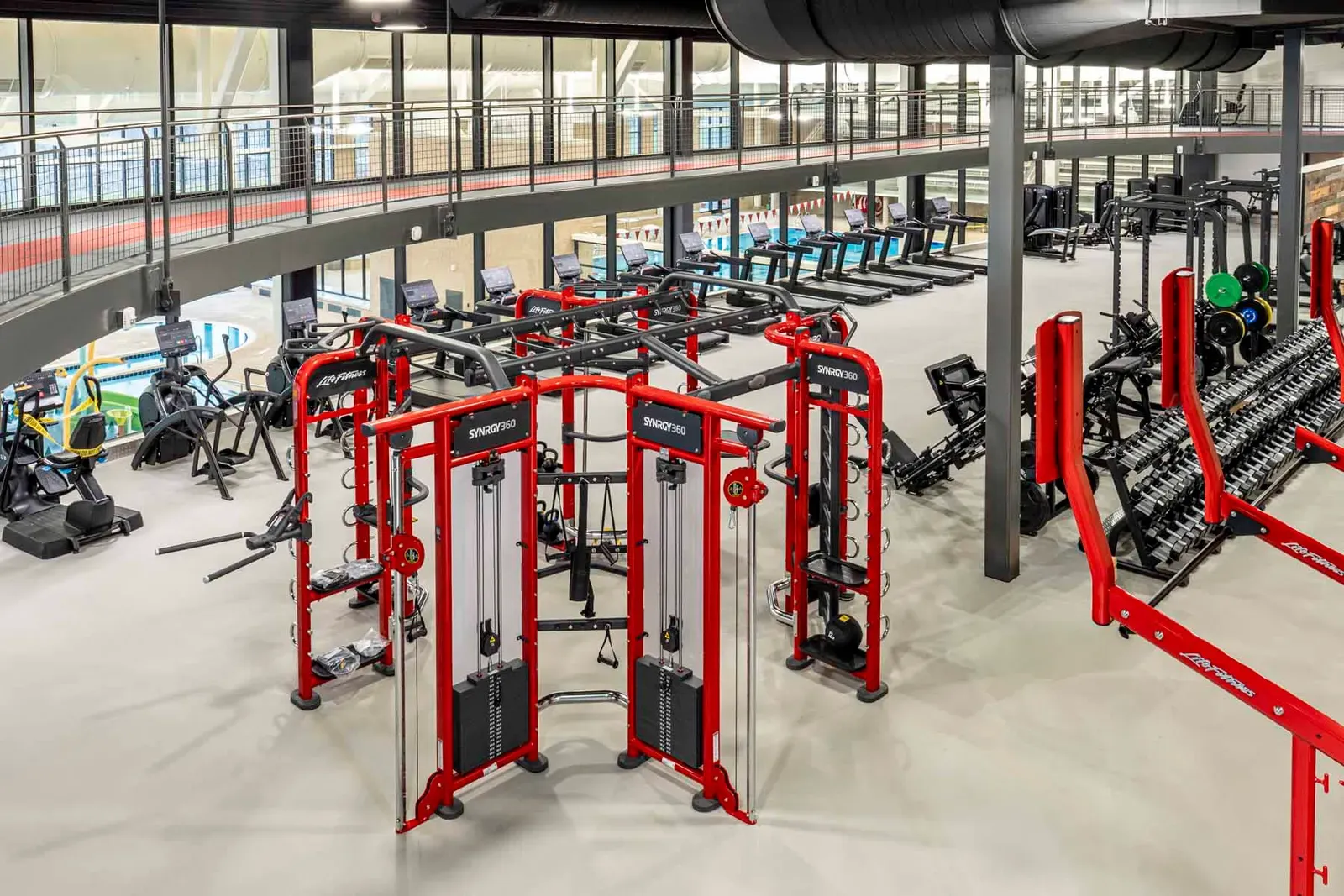 Fitness floor seen from above, with red cable machines, cardio equipment and weight racks, and a railed upper level behind.