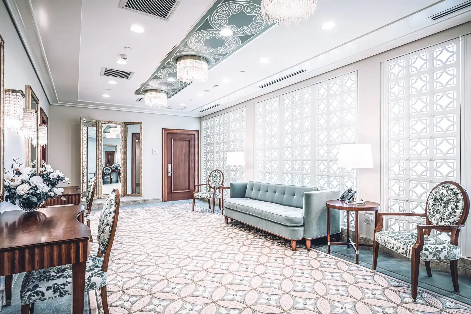 Hallway with a patterned carpet, a blue tufted sofa, upholstered side chairs and white lattice-patterned wall panels