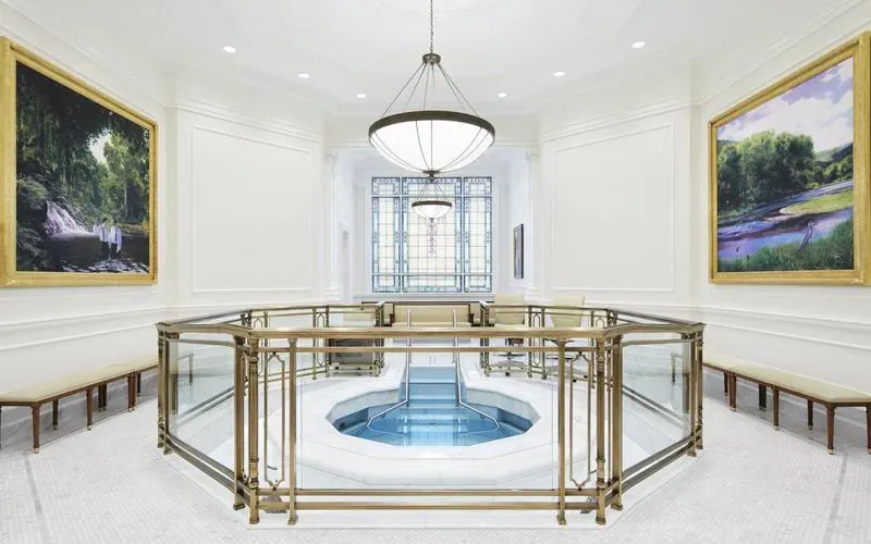 Baptistry with a font behind a glass and brass railing, a stained-glass window and large framed paintings