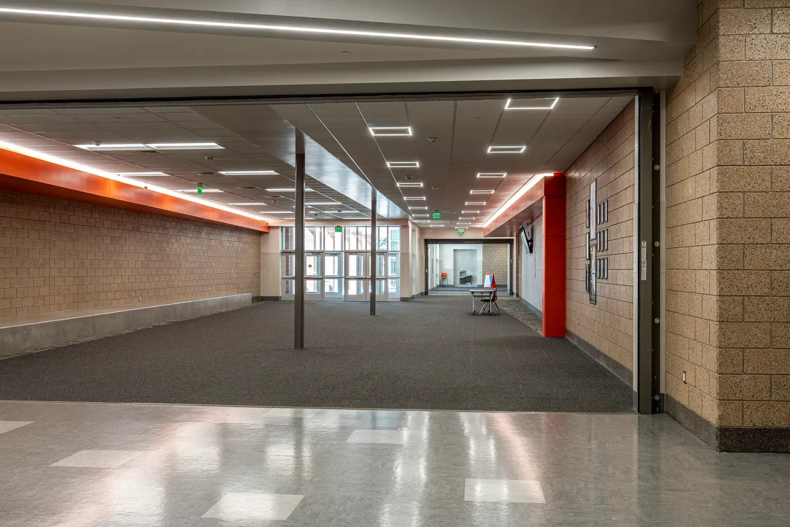 Wide corridor with tan block walls, orange accent trim and a dark carpeted opening toward glass doors.