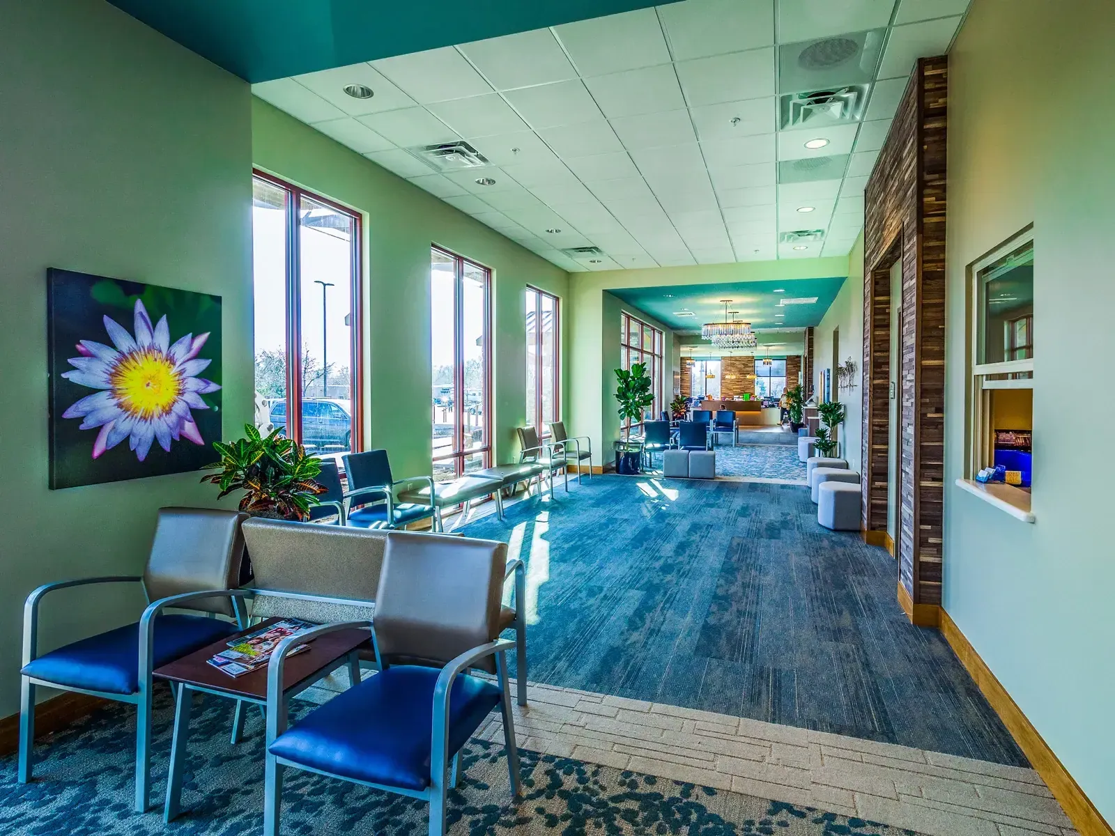 Waiting area with blue upholstered chairs, floral artwork and tall windows, opening onto a wide carpeted hall.