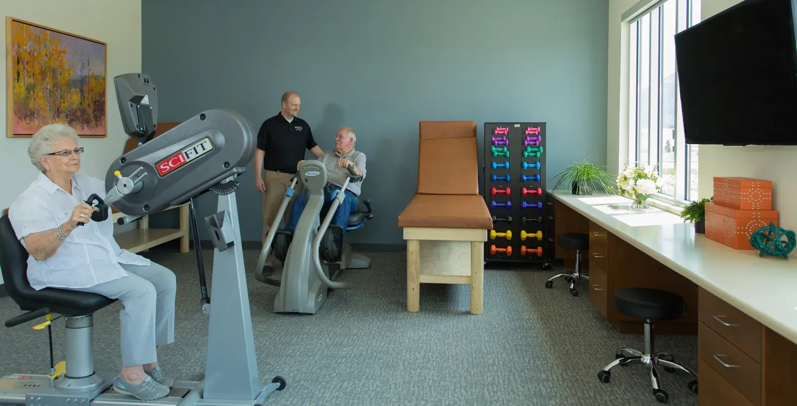 Therapy room with exercise machines, a rack of dumbbells, and three people.