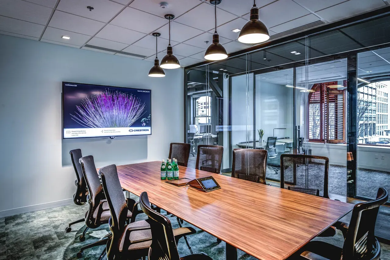Conference room with a long wood table, pendant lights, a wall-mounted display and a glass partition.