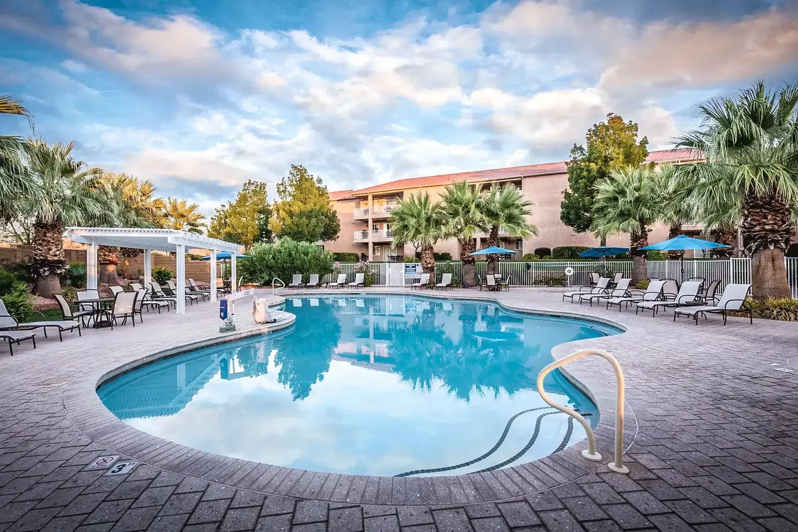 Resort swimming pool with a pergola, lounge chairs and palm trees, with a stucco building beyond