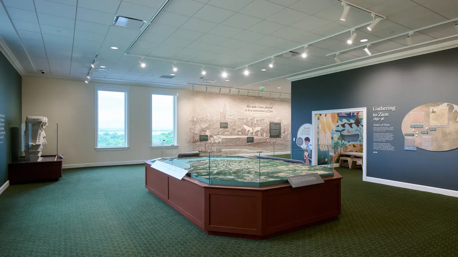 Exhibit hall with a large glass-covered relief map on a wood base, a historic mural on the wall and a display titled Gathering to Zion.