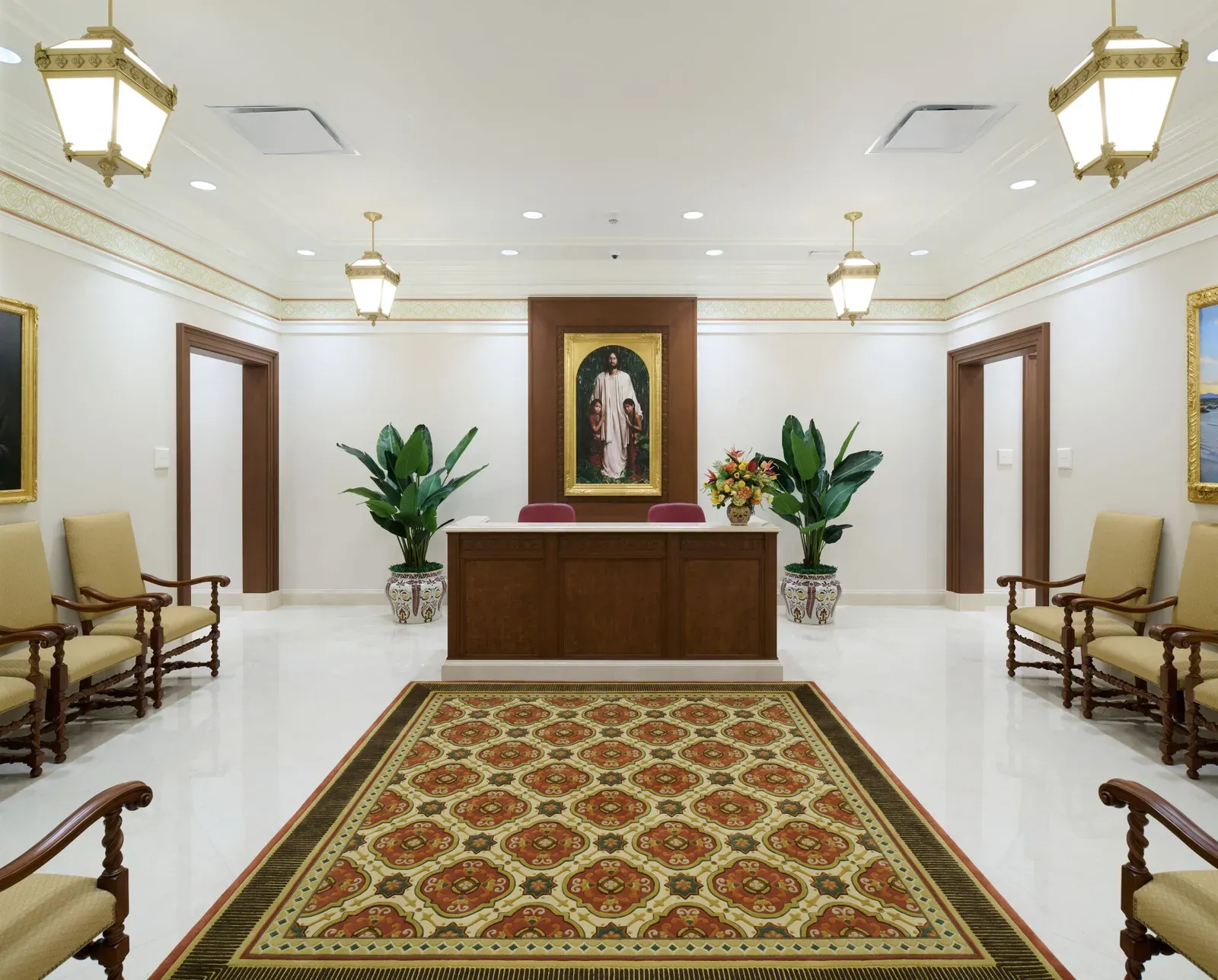 Reception area with a wood desk beneath a framed painting, patterned armchairs along the walls and a floral rug on a white floor.