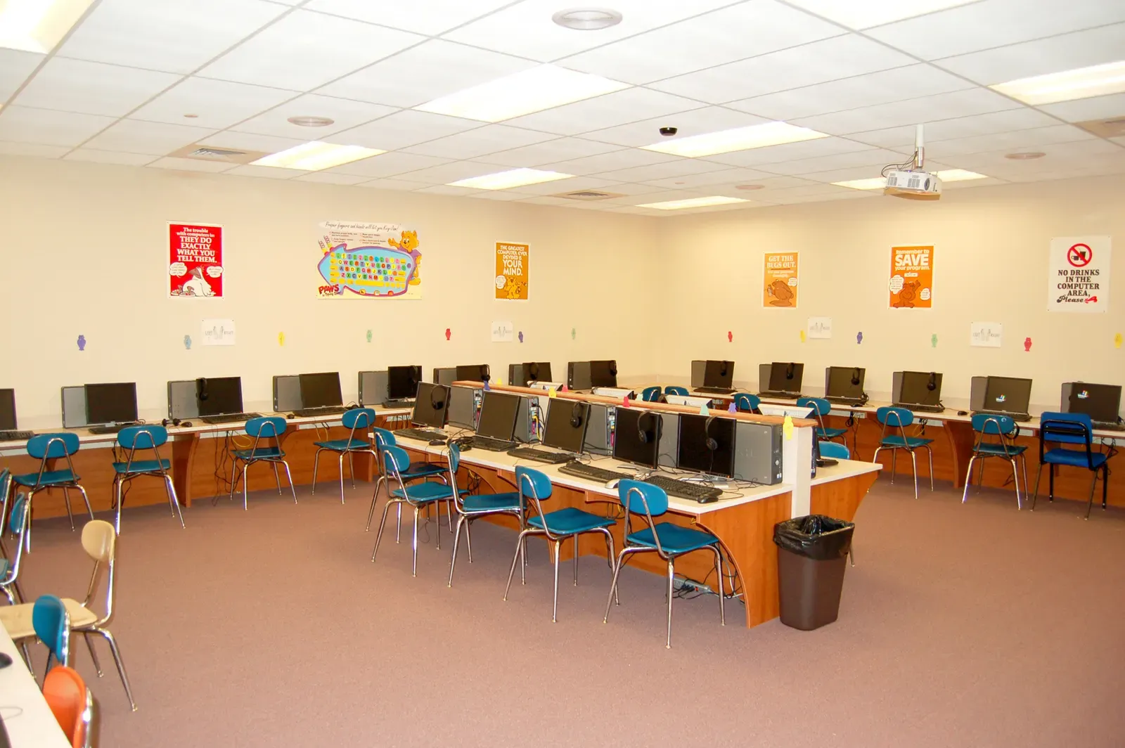 Computer lab with rows of desktop computers on long workstations, blue chairs and a ceiling projector.