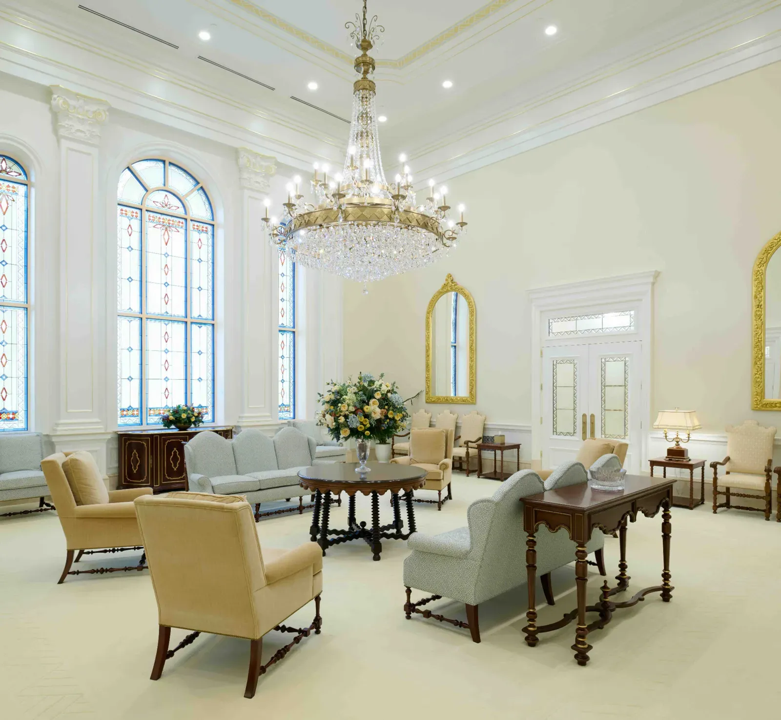 Room with a crystal chandelier, tall arched stained-glass windows, a round dark table with flowers, pale blue sofas and tan armchairs