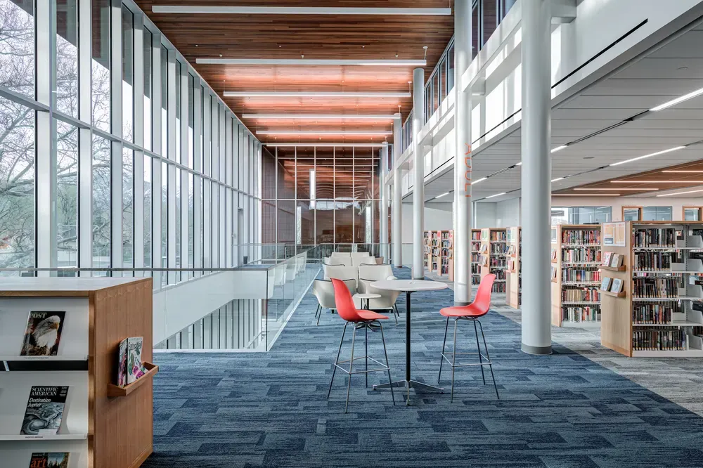 Two-story glass wall overlooking a seating area with a high table and red chairs, with bookshelves at right