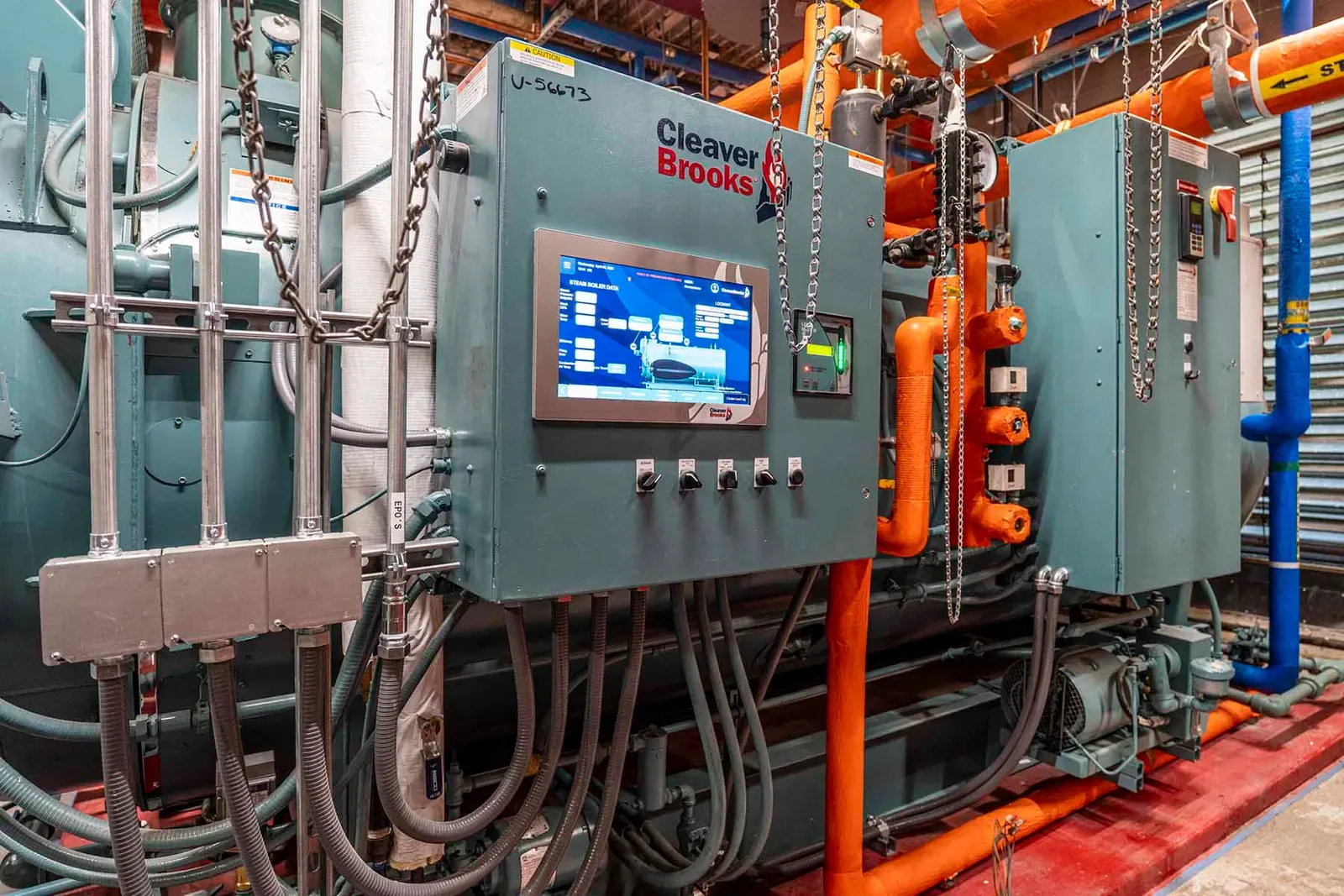 Boiler control cabinet with a touchscreen display, surrounded by orange and blue piping and conduit.