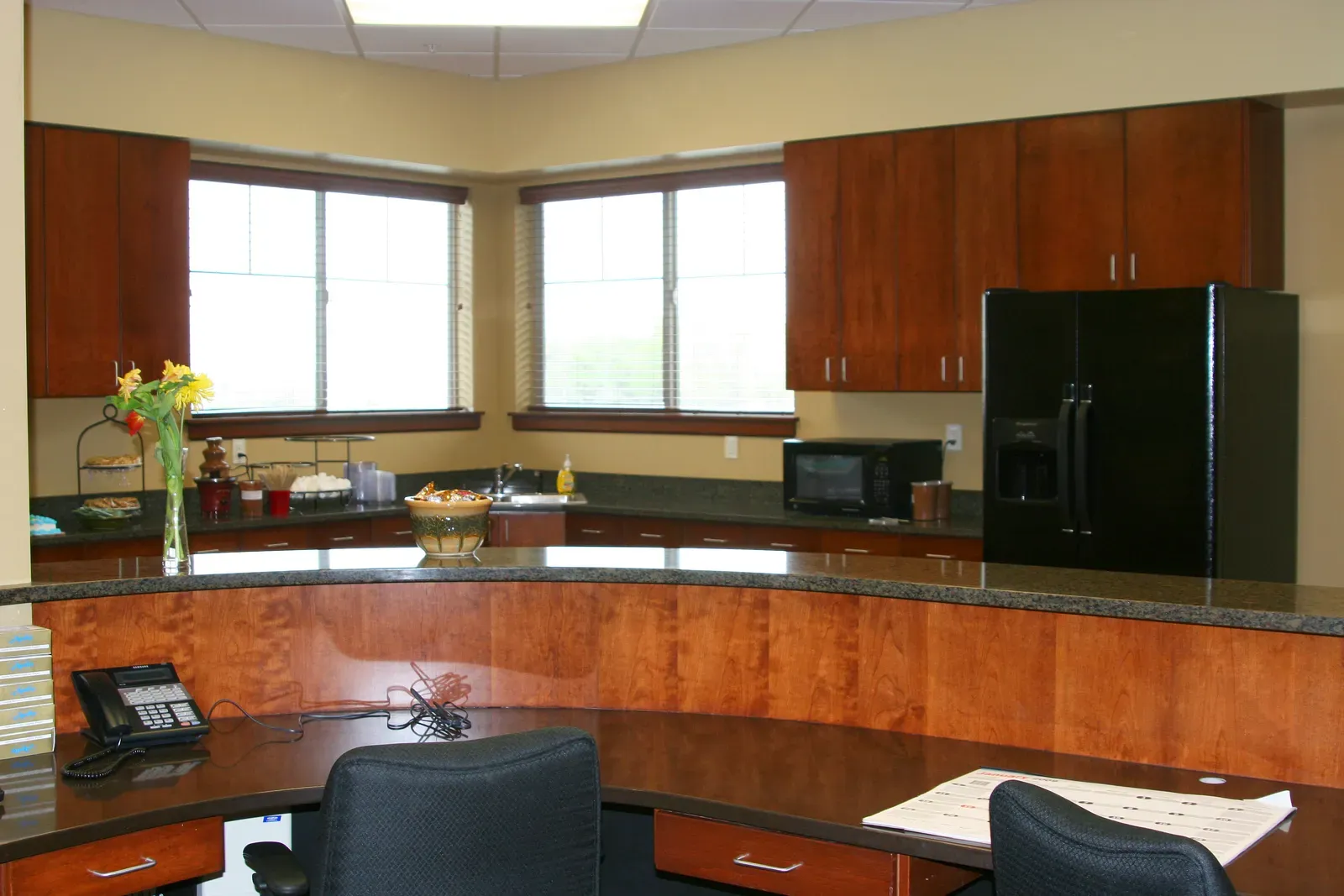 Kitchenette with cherry-colored cabinets, a black refrigerator and a curved counter in front.