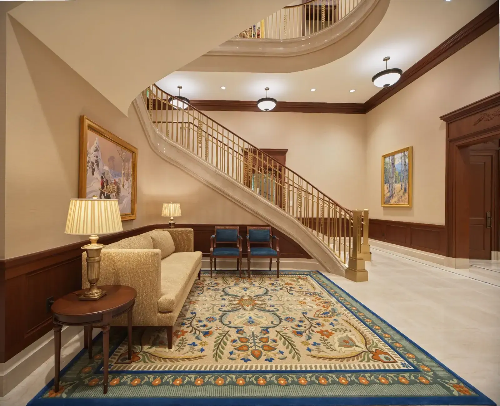 Curved stone staircase with a brass railing, a patterned rug, a sofa and two framed landscape paintings