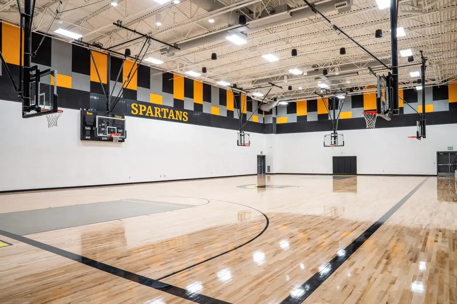 Gymnasium with a polished wood floor, ceiling-mounted hoops and black and gold sound panels along the wall.