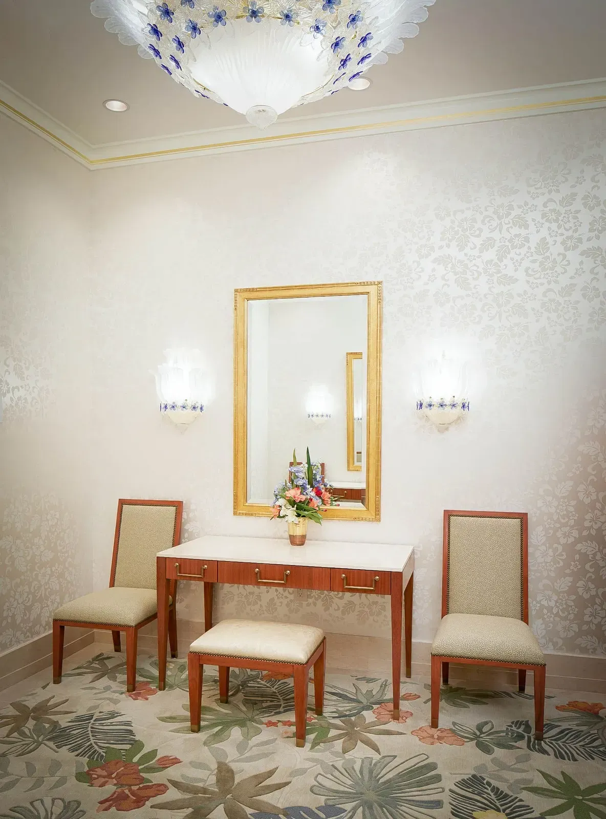 Small dressing area with a gold-framed mirror, a wood table with stools, and a flower-patterned rug.