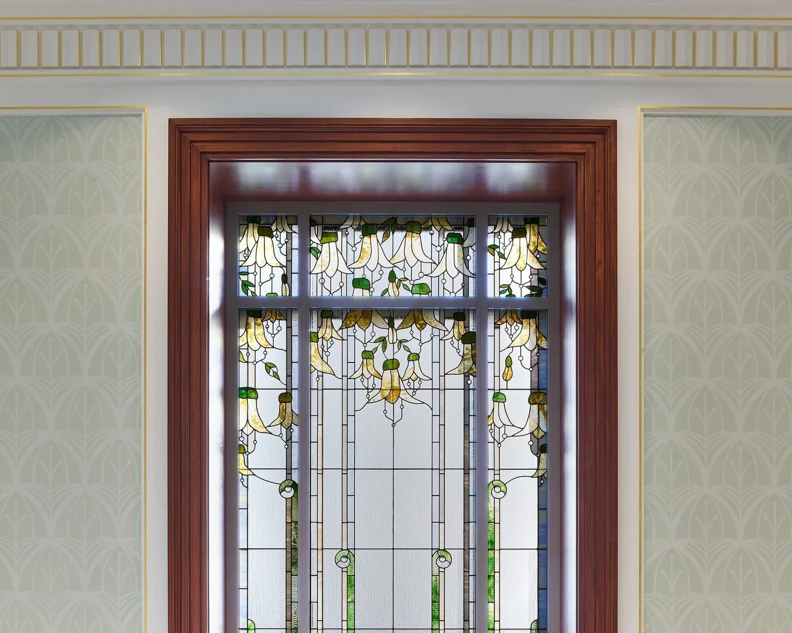 Art glass window with yellow hanging flowers and green leaves in a wood frame, set in a patterned wall panel