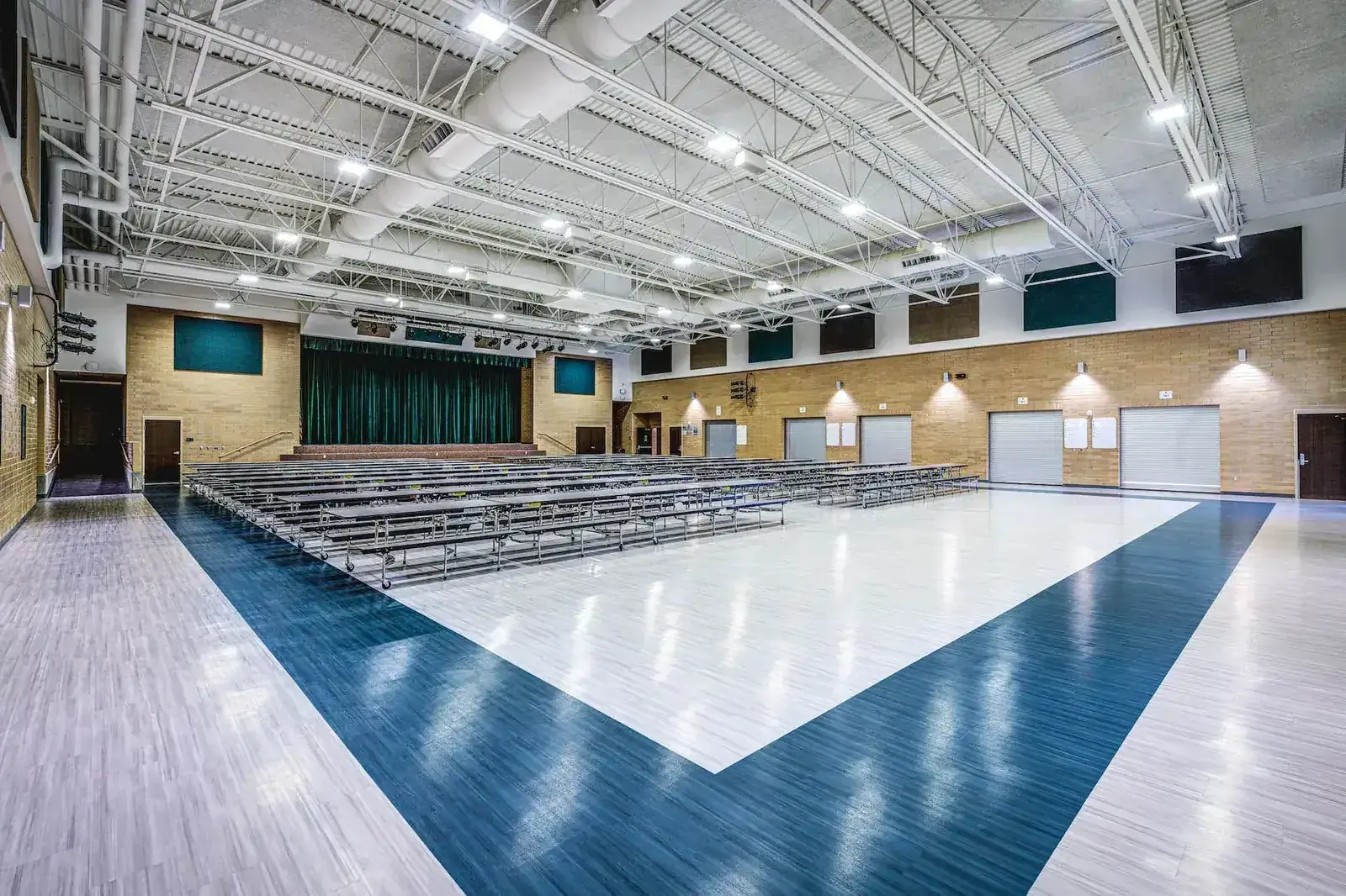 Gymnasium with a blue and white floor, folded bleachers along one side, a green curtain at the far end and exposed steel trusses overhead.