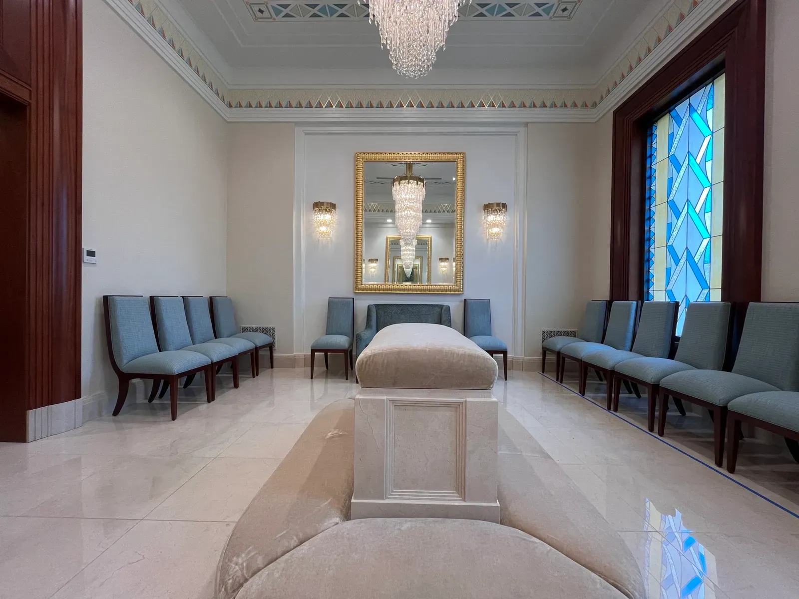 Room with an upholstered altar at the center, a crystal chandelier, a gold-framed mirror and blue chairs along the walls.