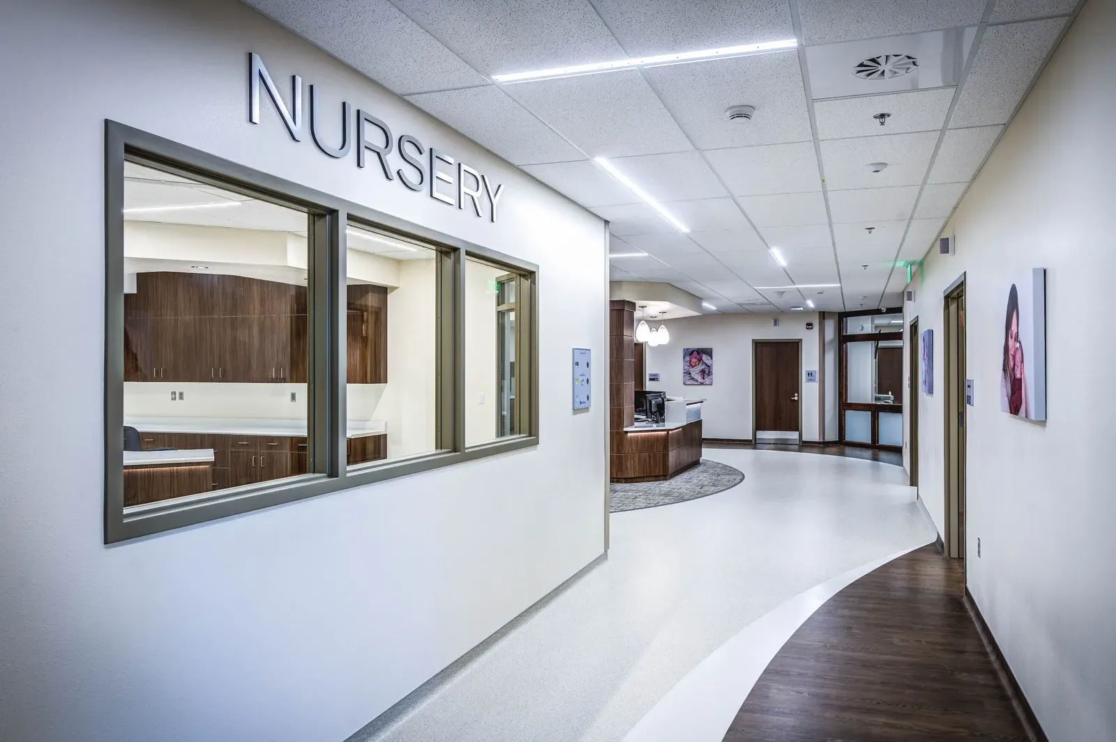 Corridor with a window labeled Nursery and a curved flooring pattern leading to a nurses' desk
