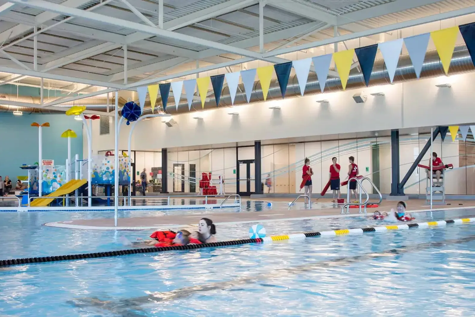 Recreation pool with lifeguards in red, a small slide and play structure, and pennants hanging from the roof.