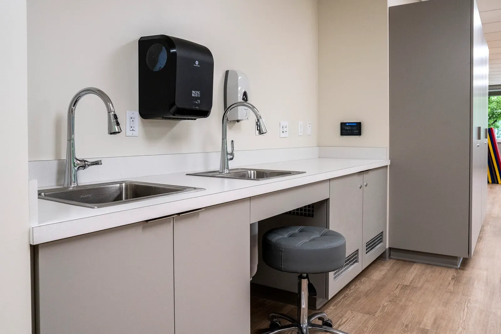 Counter with two stainless sinks, a paper towel dispenser, gray cabinets and a rolling stool.