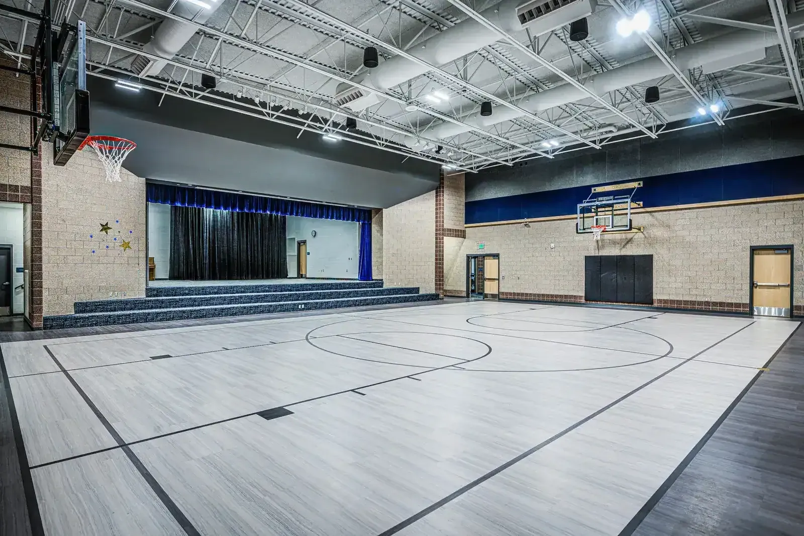Gymnasium with a wood-look floor, basketball hoops and a stage with a blue curtain at one end.