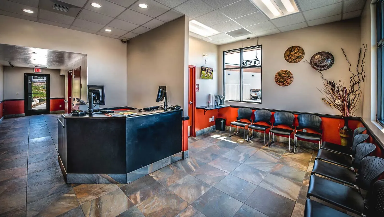 Waiting room with a dark reception counter, tile floor and a row of chairs along a window.