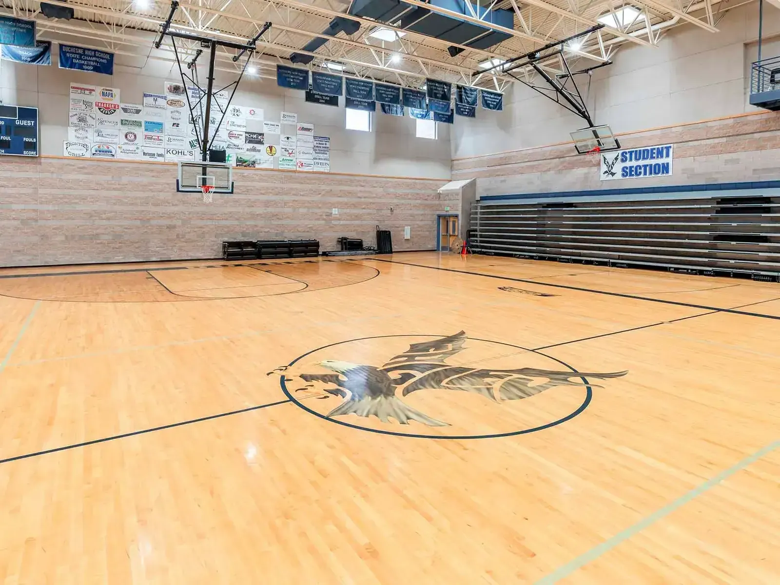 Gymnasium with a wood floor, an eagle logo at center court, and bleachers folded against the wall