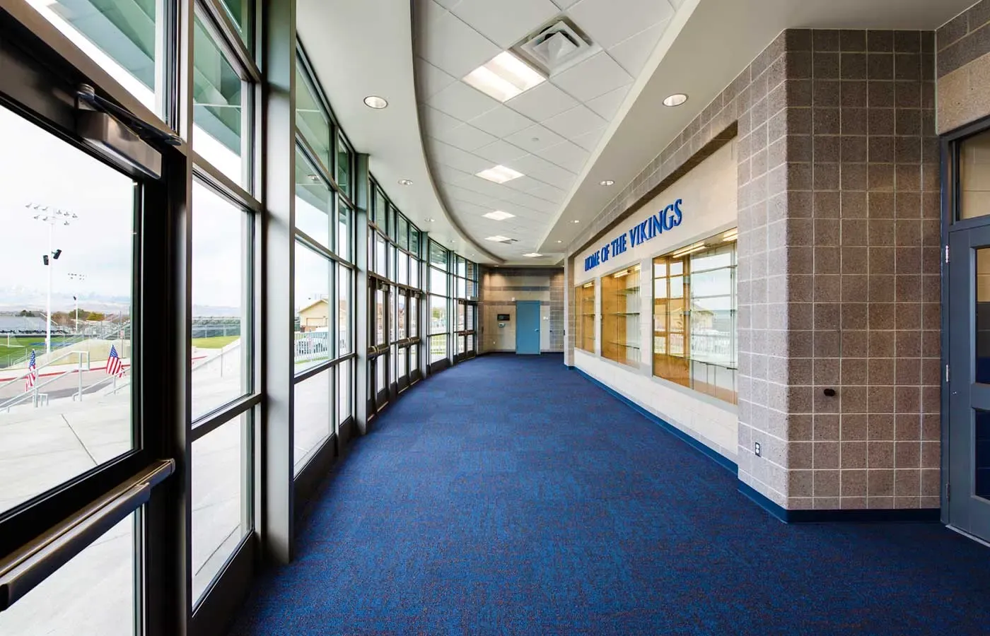 Curved corridor with tall windows, blue carpet and a "Home of the Vikings" sign above a display case.