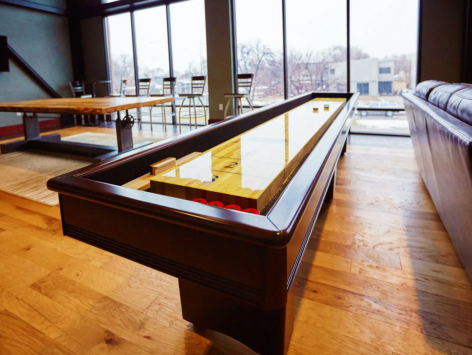 Shuffleboard table in front of a tall window wall, with a long wood table and bar stools behind.