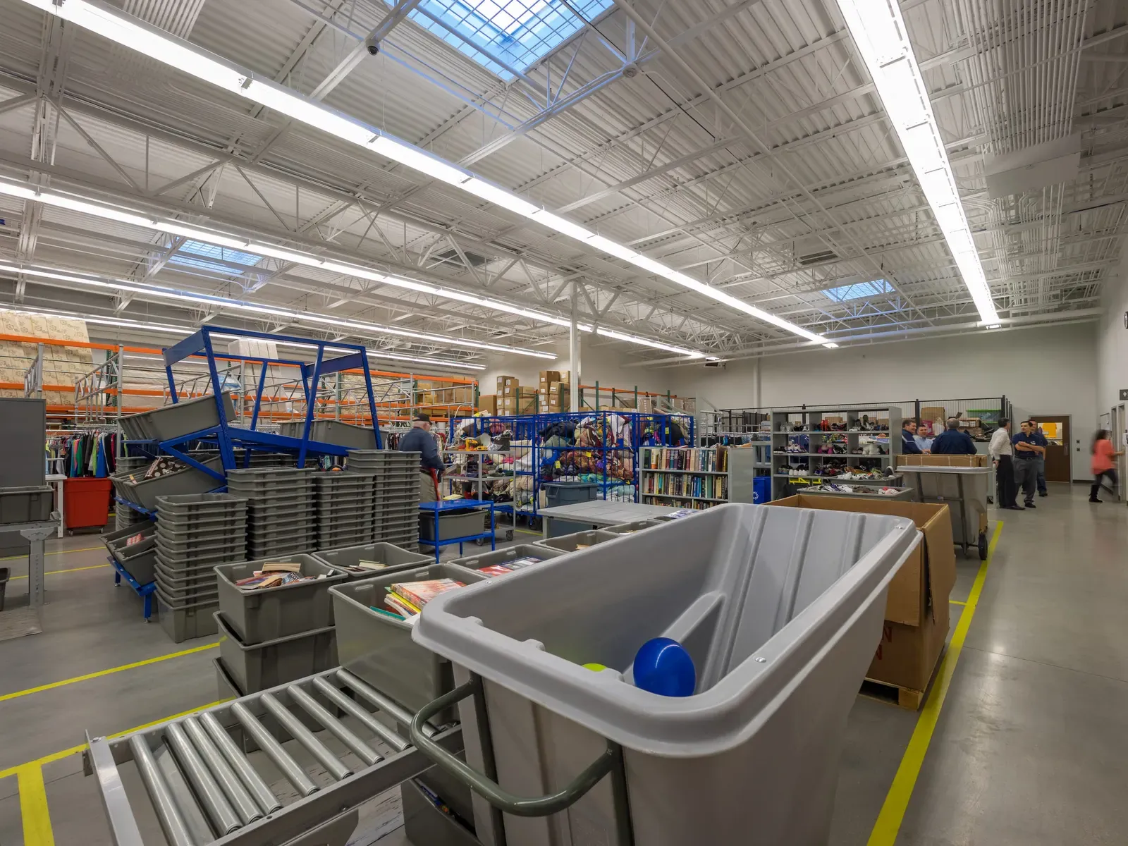 Back-of-house processing area with gray bins and rolling carts under a high steel-truss ceiling with skylights.