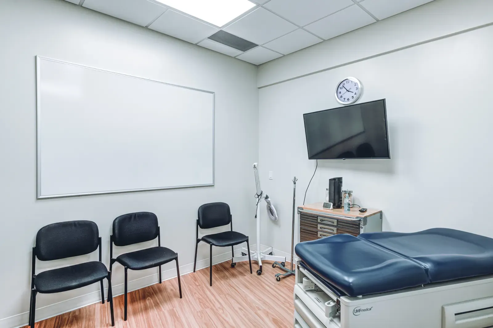Exam-style training room with a padded exam table, three chairs, a whiteboard and a wall-mounted screen.