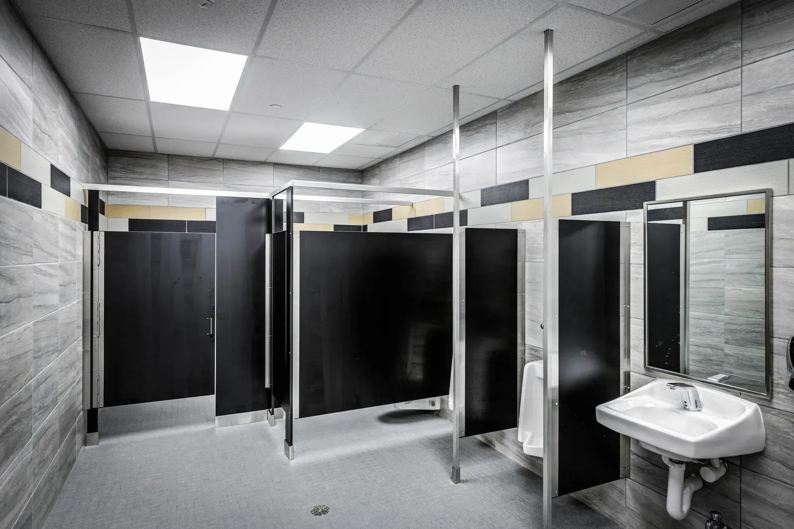 Restroom with dark stall partitions, gray stone-look wall tile and a wall-mounted sink
