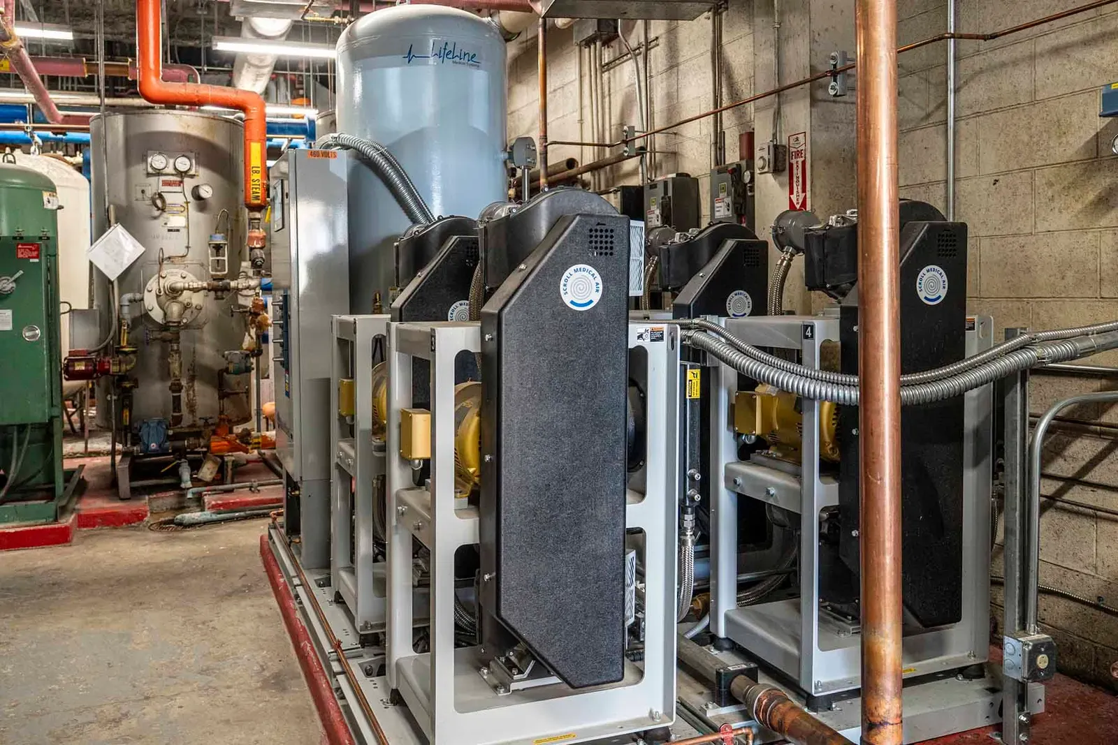 Mechanical room equipment on steel frames with a gray tank behind, copper and orange piping overhead.