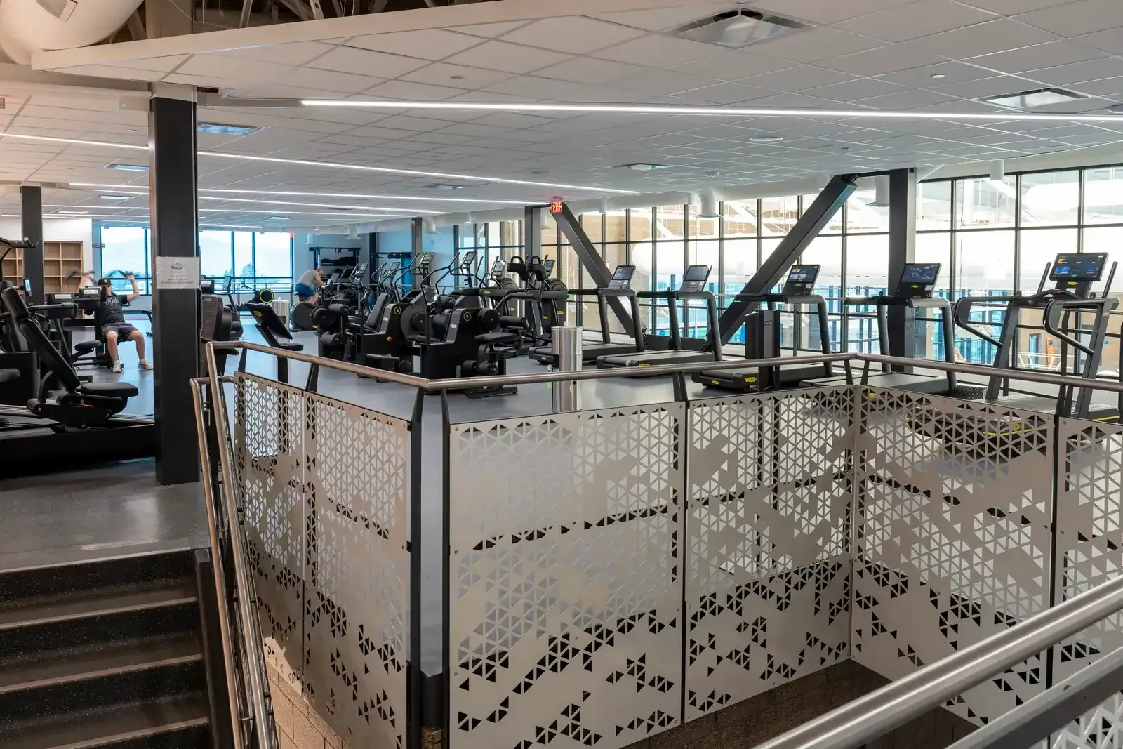 Fitness area with cardio machines beside windows, behind a perforated metal railing at the top of a stair.