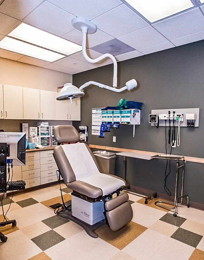 Exam room with a padded exam chair, an overhead exam light, wall-mounted diagnostic instruments and wood cabinets