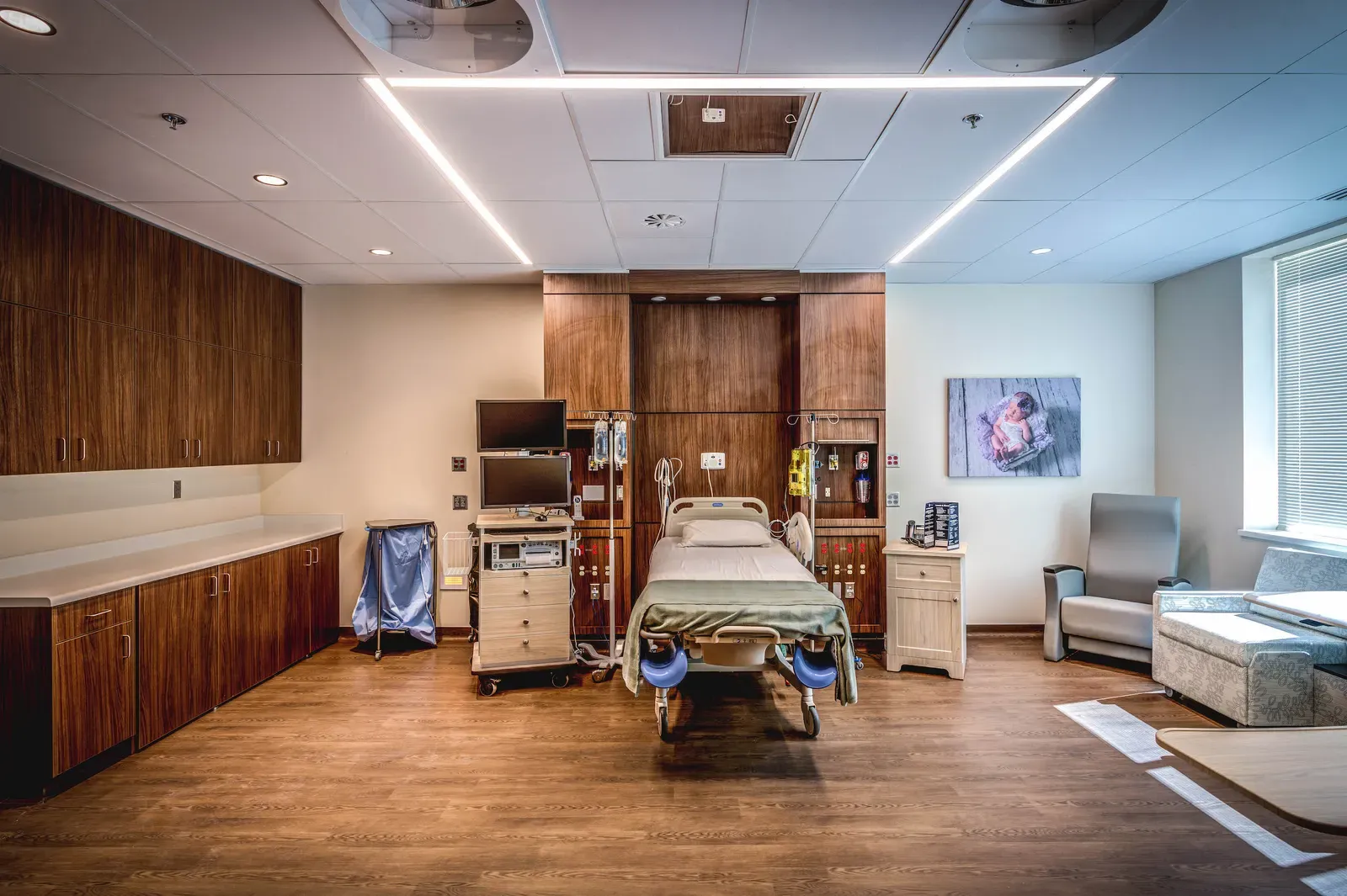 Patient room with a hospital bed, wood headwall and cabinetry, and a recliner and sofa by the window