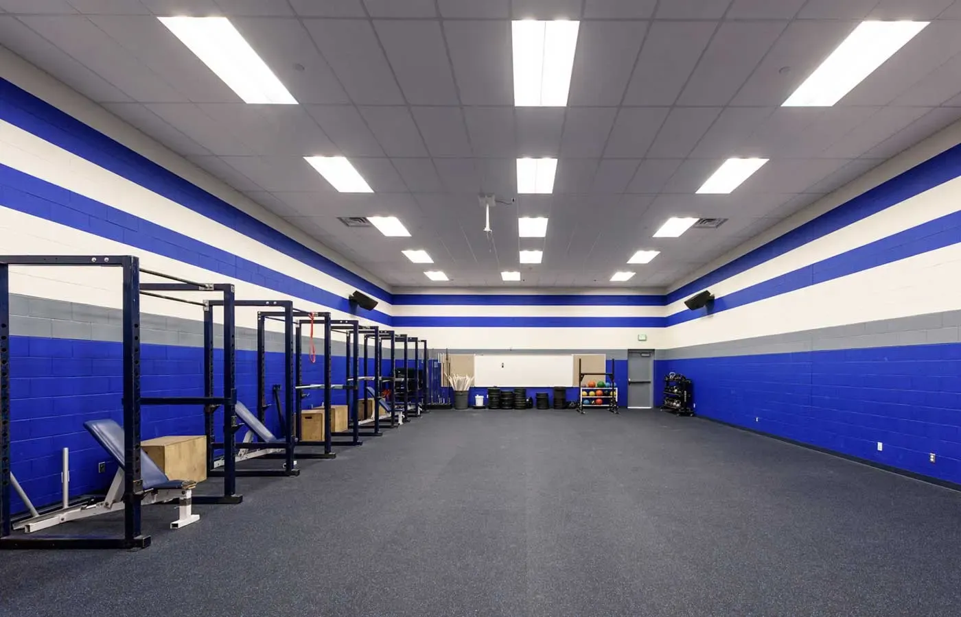 Weight room with squat racks, blue and white striped block walls and a dark rubber floor.