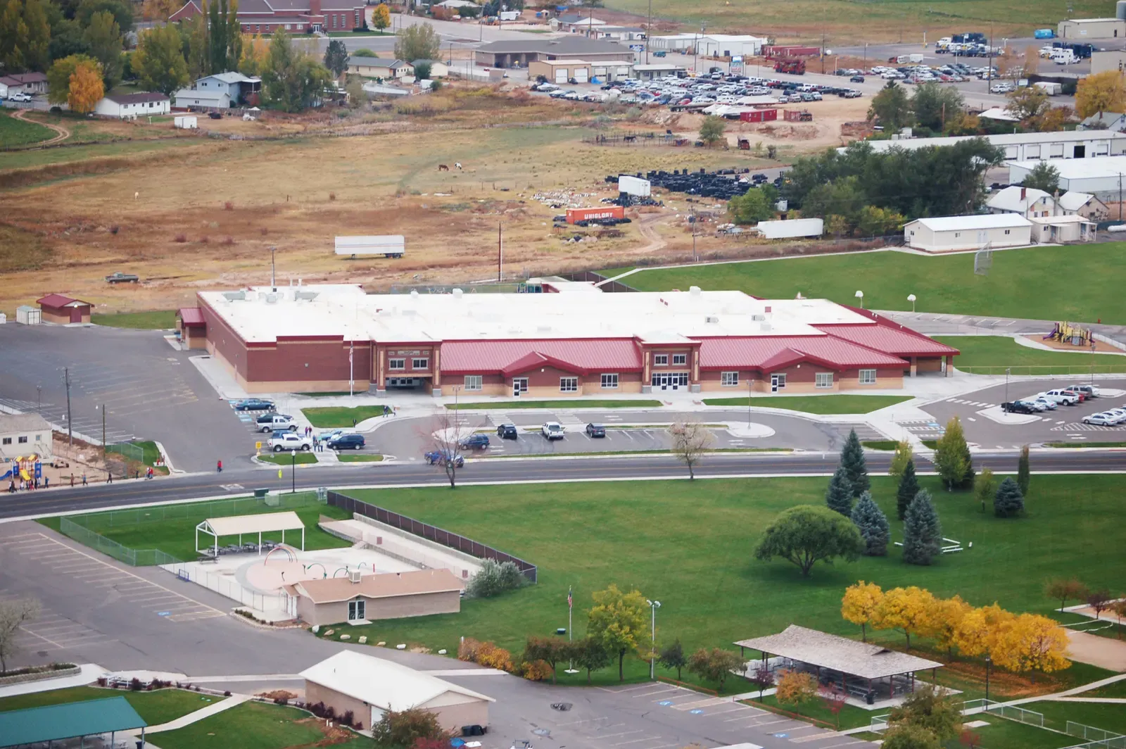 Aerial view of the school with its parking lot and grounds, trees in autumn color and open land beyond.