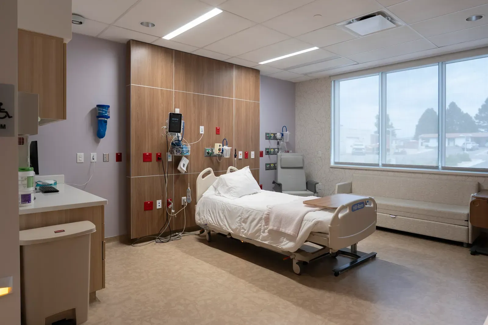 Patient room with a hospital bed, a wood-panel headwall, a recliner and a window bench.