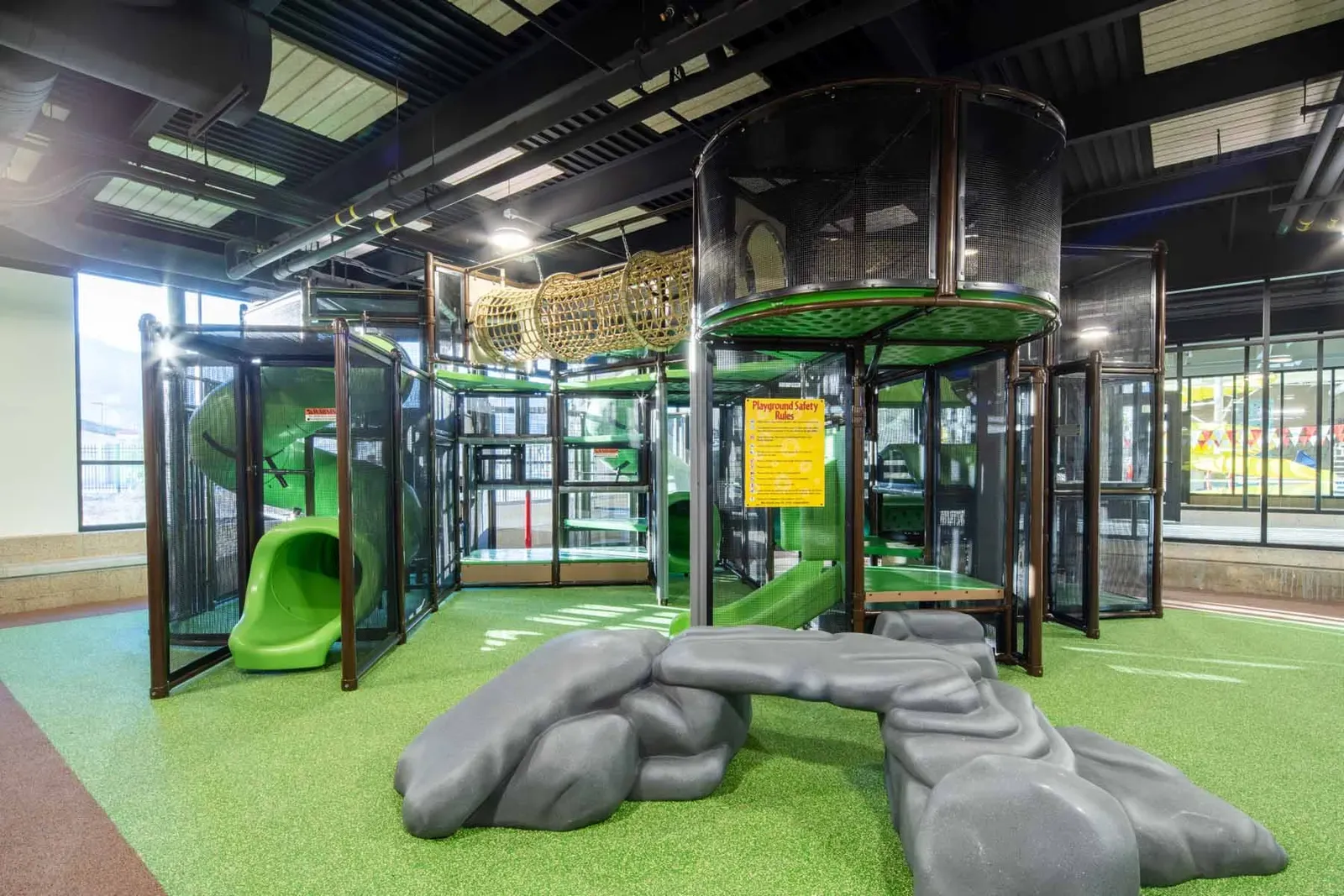 Indoor play area with an enclosed multi-level play structure, a green slide and gray climbing rocks on green turf flooring.