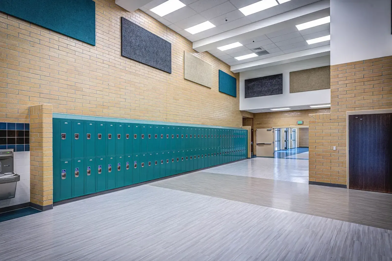 Corridor with a long row of teal lockers, brick walls and sound-absorbing wall panels.