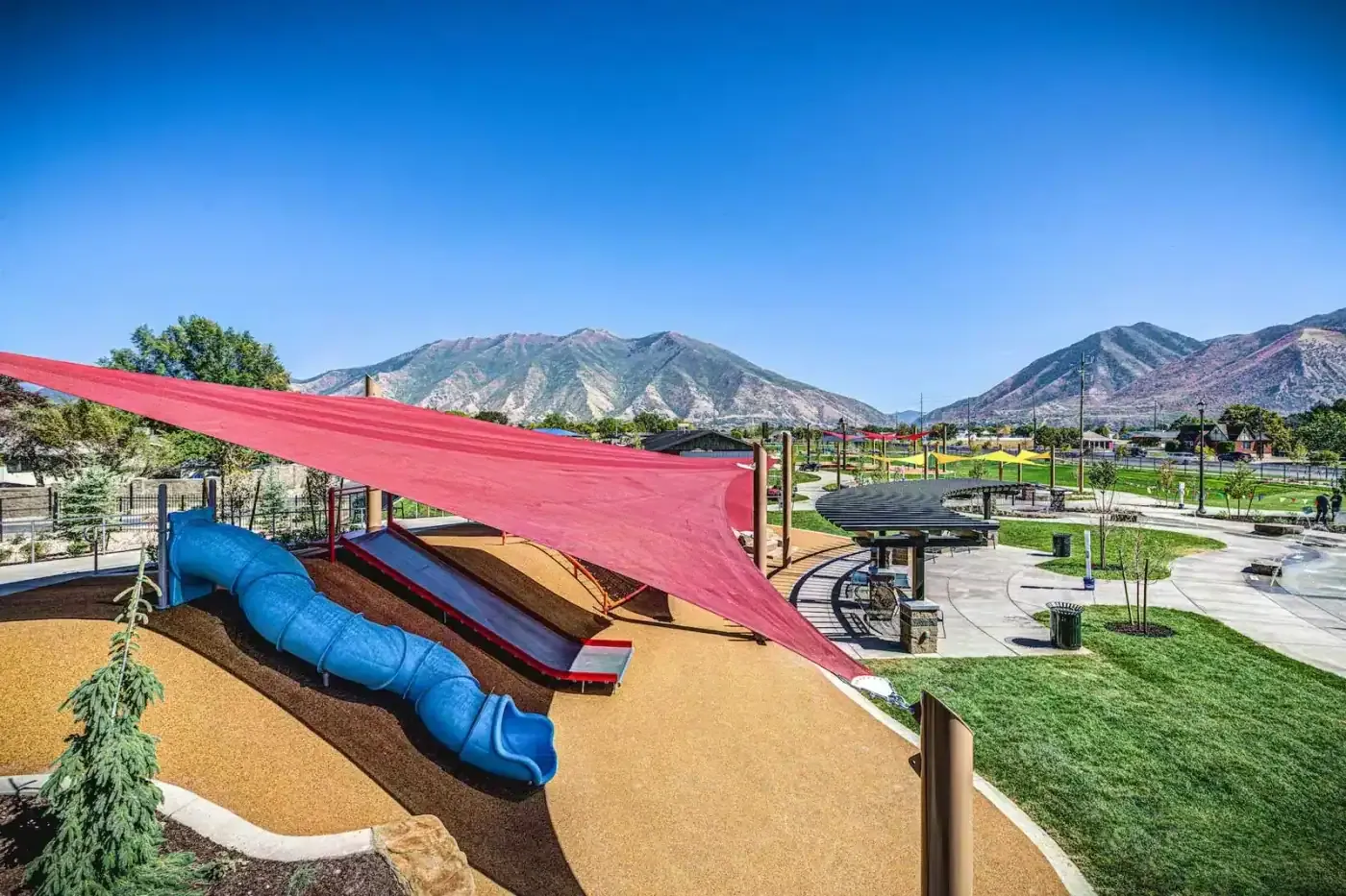Blue tube slide and red slide on a rubber-surfaced mound under a red shade sail, with mountains beyond