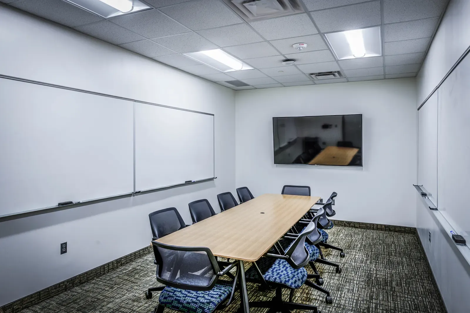 Small meeting room with a long table, a wall-mounted display, and whiteboards