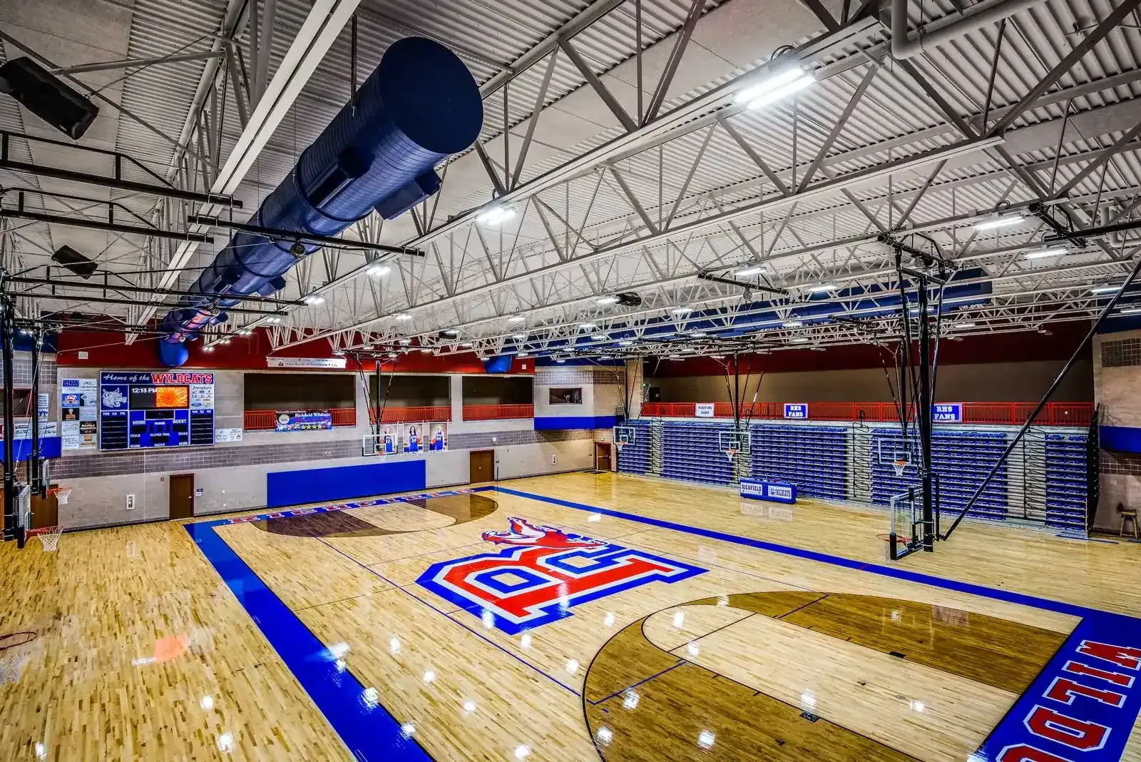 Gymnasium with a wood floor, a large painted school logo, bleachers and an exposed steel roof structure