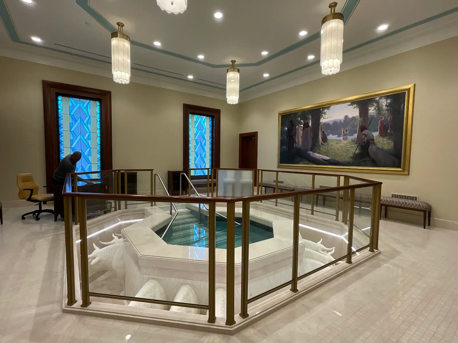 Baptismal font with a glass and brass railing, sculpted white oxen beneath it, blue art glass windows and a large framed painting of a wooded river scene.