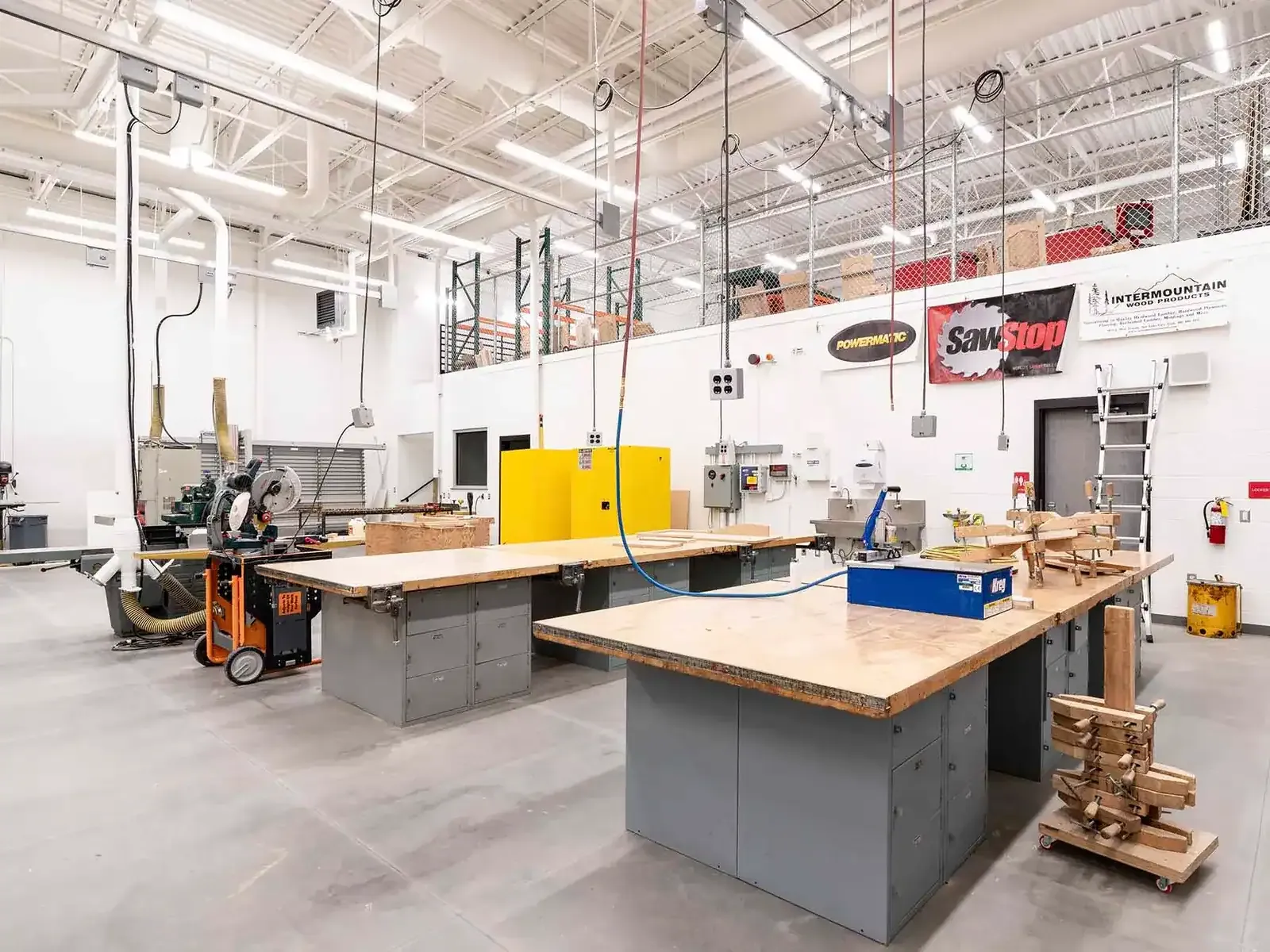 Career and technical education woodshop with workbenches, a miter saw, and dust collection piping overhead.