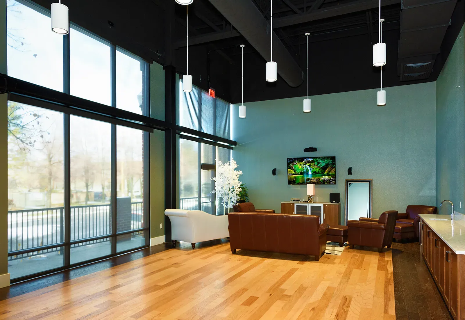 Lounge with leather seating, a wall-mounted screen, pendant lights and tall windows over a wood floor.