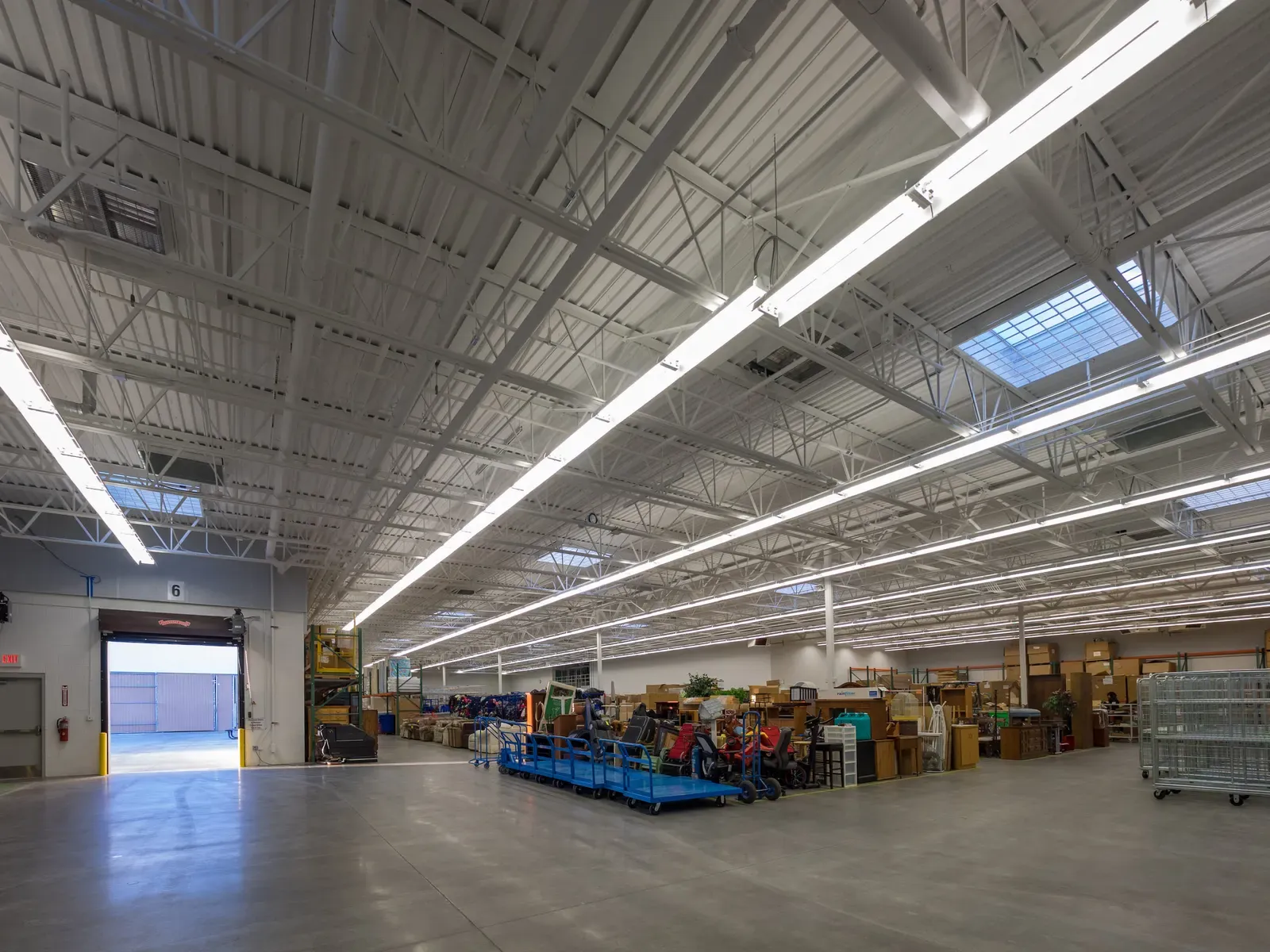 Warehouse area with a high steel ceiling, skylights, a loading dock door at left and carts of goods in the distance.
